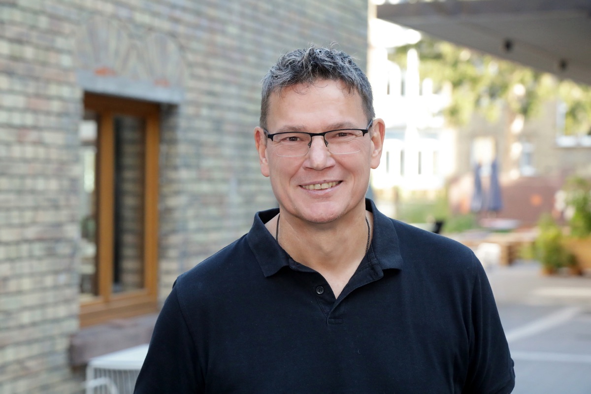 Ralph Suikat, lächelnd, mit Brille und dunklem Poloshirt vor einer Backsteinwand